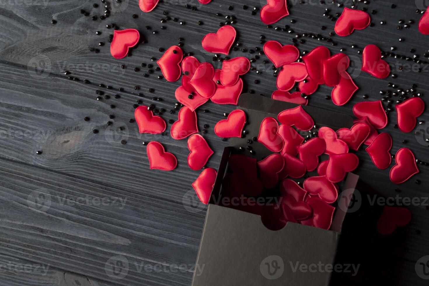 röd hjärtan är hällde ut av en svart gåva låda på en mörk blå trä- tabell. foto
