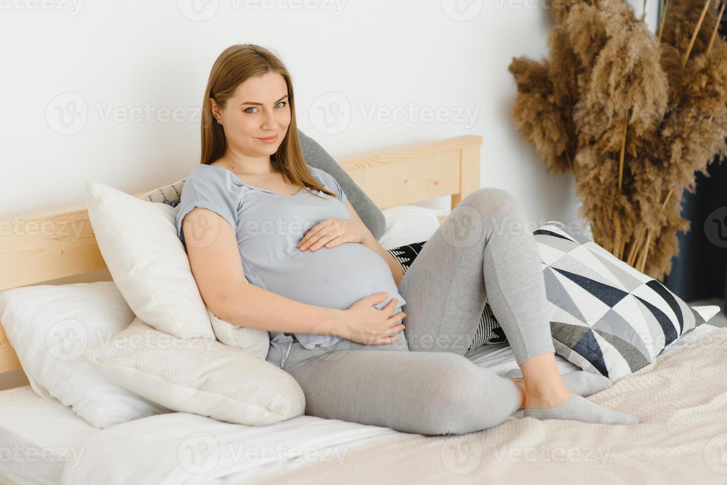 gravid ung europeisk kvinna Sammanträde på en stor säng och strök henne stor mage, 7 månad graviditet, väntar för de födelse av henne bebis. foto