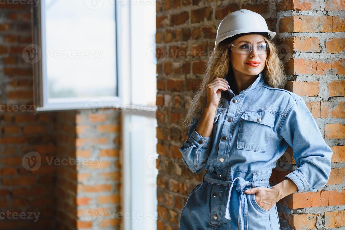 attraktiv kvinna konstruktion arbetstagare i hjälm. självsäker ung specialist i rutig blå skjorta i jeans stående i tömma rum. interiör design och renovering service. foto