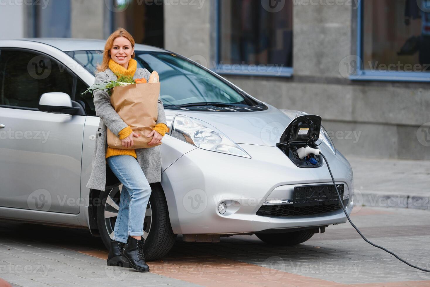 kvinna med en mobil telefon nära laddning elektrisk bil. fordon laddning på offentlig laddning station utomhus. bil delning begrepp foto