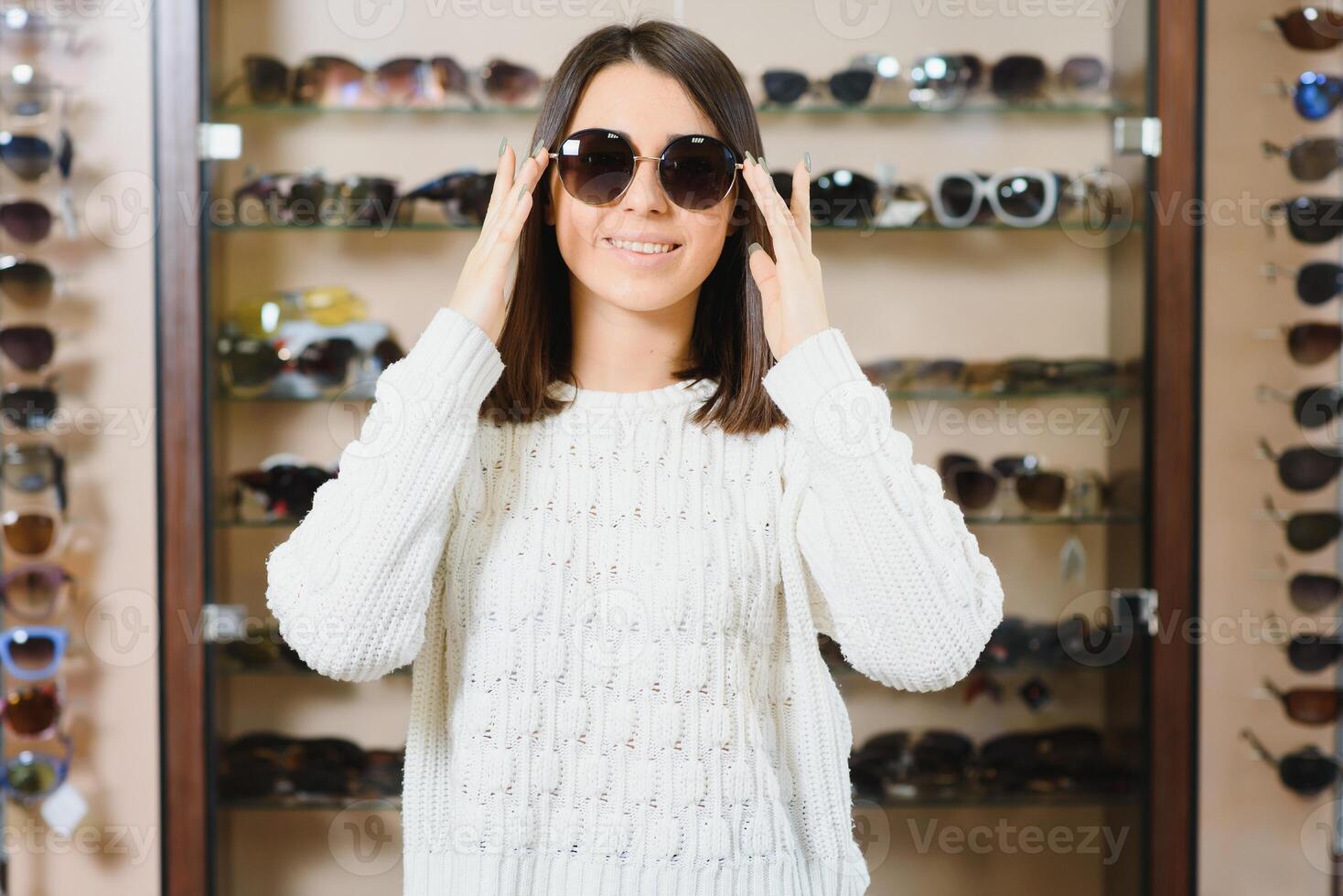 som ett är bättre passa mig. porträtt av glad tvekande kvinna i optiker Lagra, framställning beslut, innehav eleganta solglasögon, välja Vad hon skall köpa. bra rabatter för uppköp två par. foto