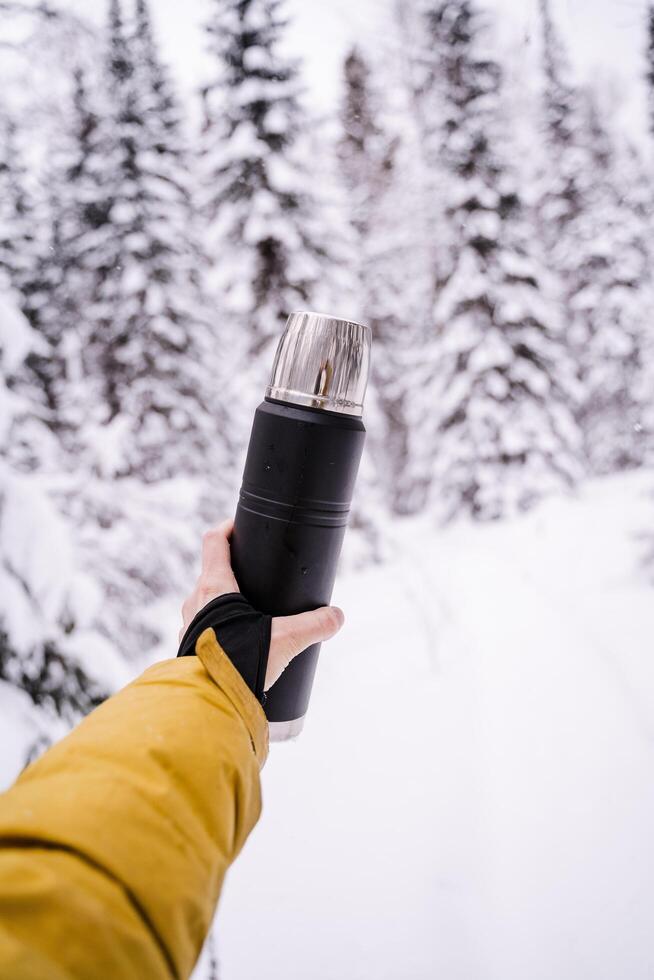 svart matt termos i hand. metall lock och mugg.drink värma te och kaffe från en termos på en promenad i vinter. snötäckt skog på en tyst dag. ljus kläder foto