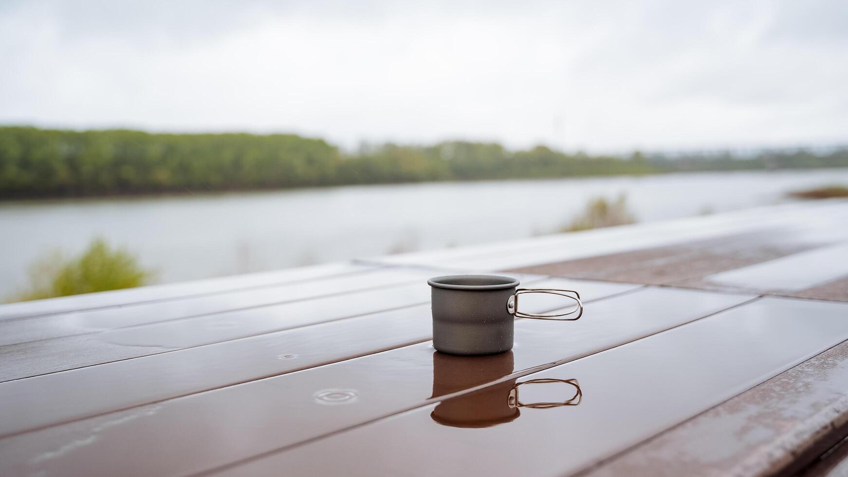 kaffe råna i natur, camping redskap, dricka varm te på en vandra, reflekterande de silhuett av en glas i en pöl av vatten, en kopp stående på de tabell våt, regn i de skog foto