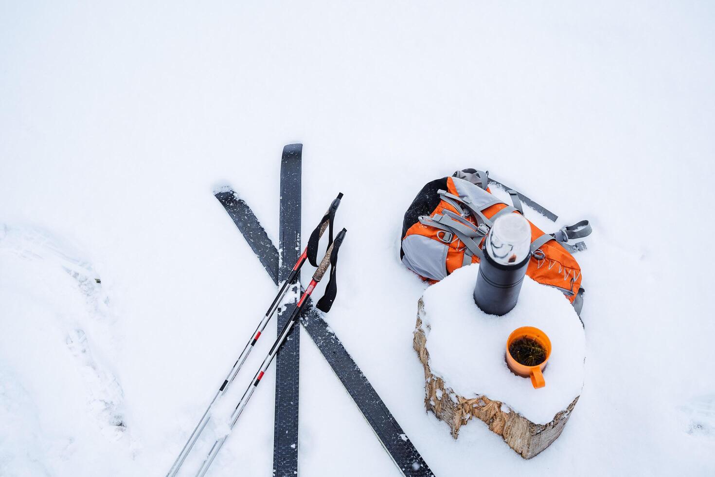 falsk poler och skidor lögn på de snö, Nästa till en stubbe med en termos, orange vandring ryggsäck och en råna av te. vinter- fortfarande liv i de snö. skidåkning i de skog, färsk luft foto