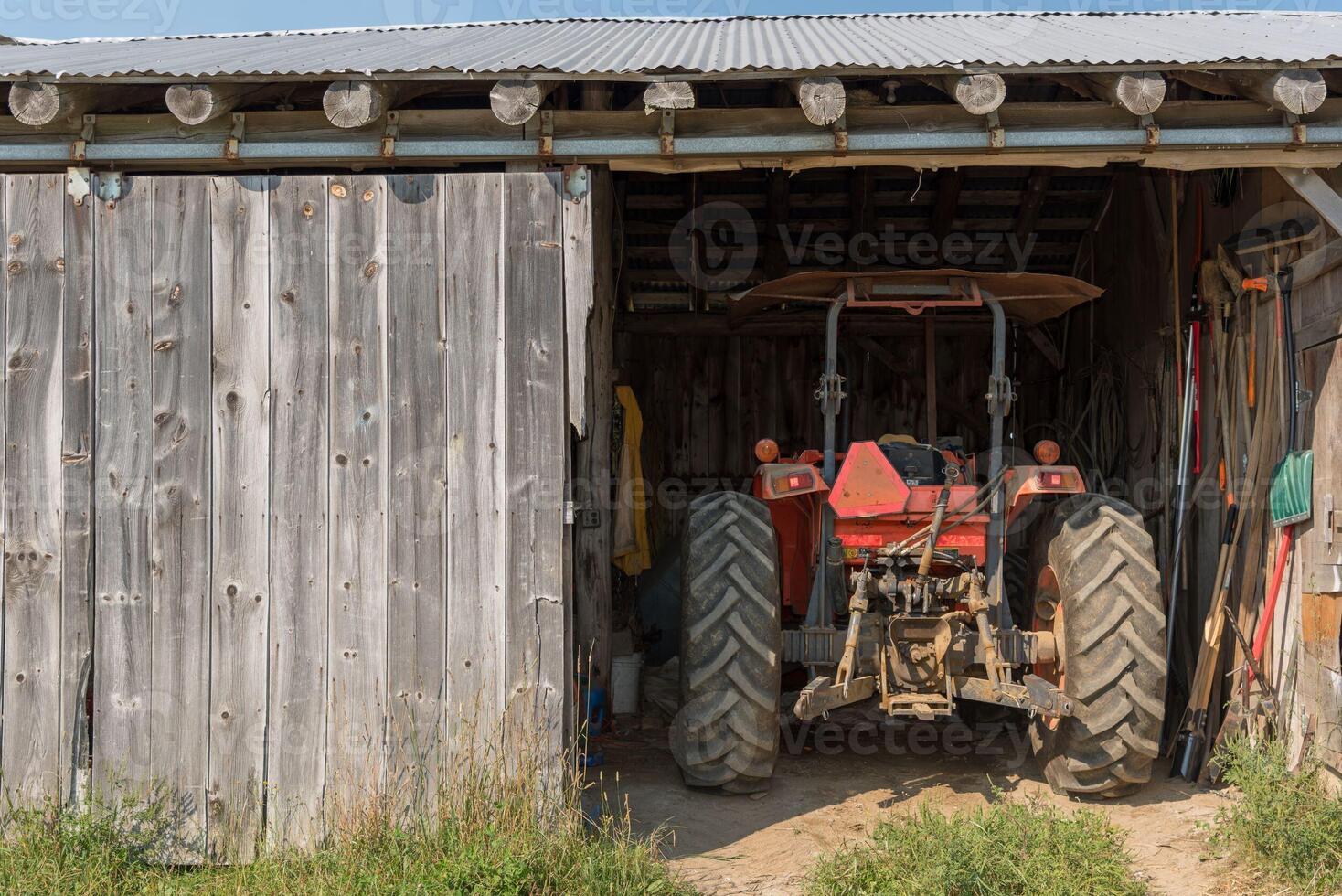 traktor parkerad i en skjul med bruka verktyg och Utrustning på en solig sent eftermiddag foto