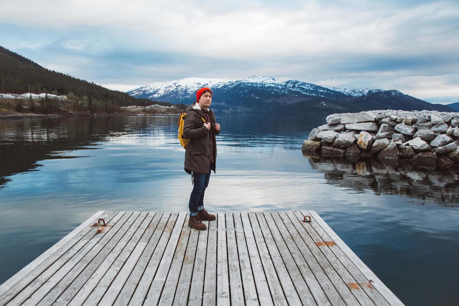 resenär man med en gul ryggsäck bär en röd hatt som står på bakgrunden av berg och sjö träbrygga. resa livsstilskoncept foto