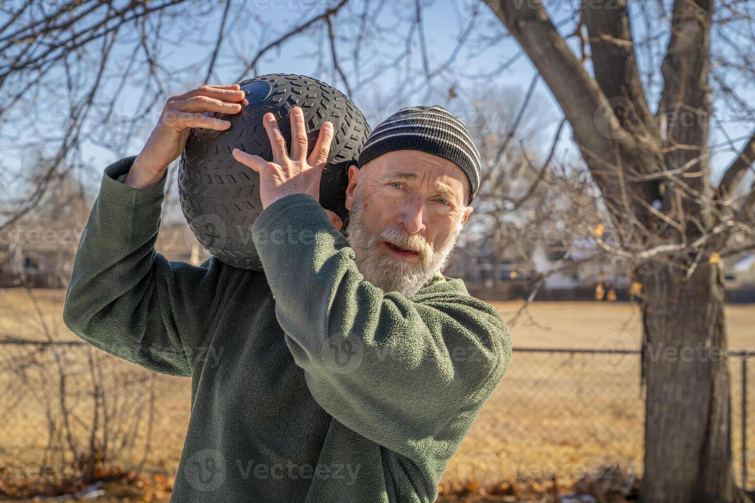 senior man är utövar med en tung, 50 lb, slam boll i hans bakgård, solig vinter- dag foto