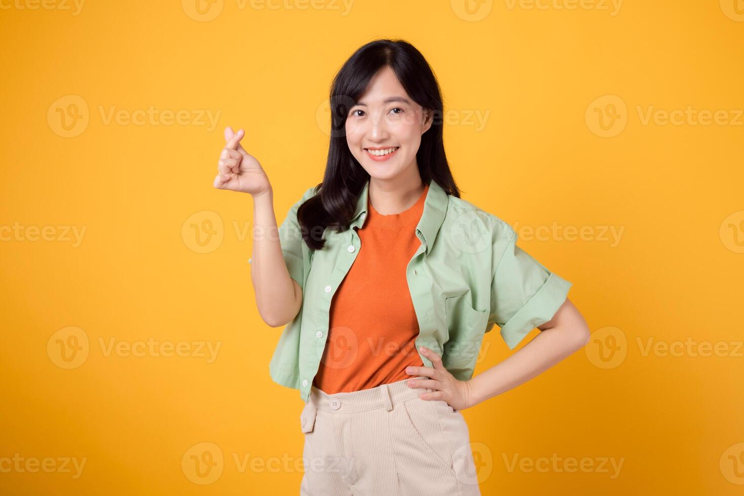 lycka med en ung asiatisk kvinna i henne 30-talet, klädd i ett orange skjorta och grön hoppare. henne mini hjärta gest, höft håll, och mild leende framföra en djupgående meddelande genom kropp språk. foto
