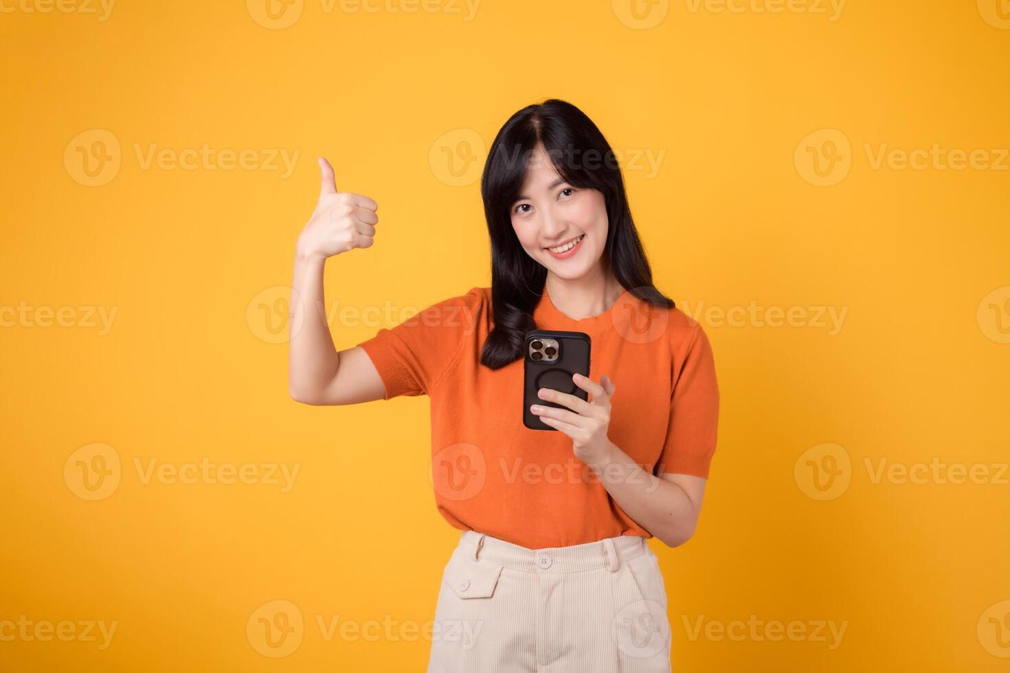 vibrerande asiatisk kvinna i henne 30-talet, bär orange skjorta, använder sig av smartphone med tumme upp hand tecken på vibrerande gul bakgrund. ny mobil app begrepp. foto