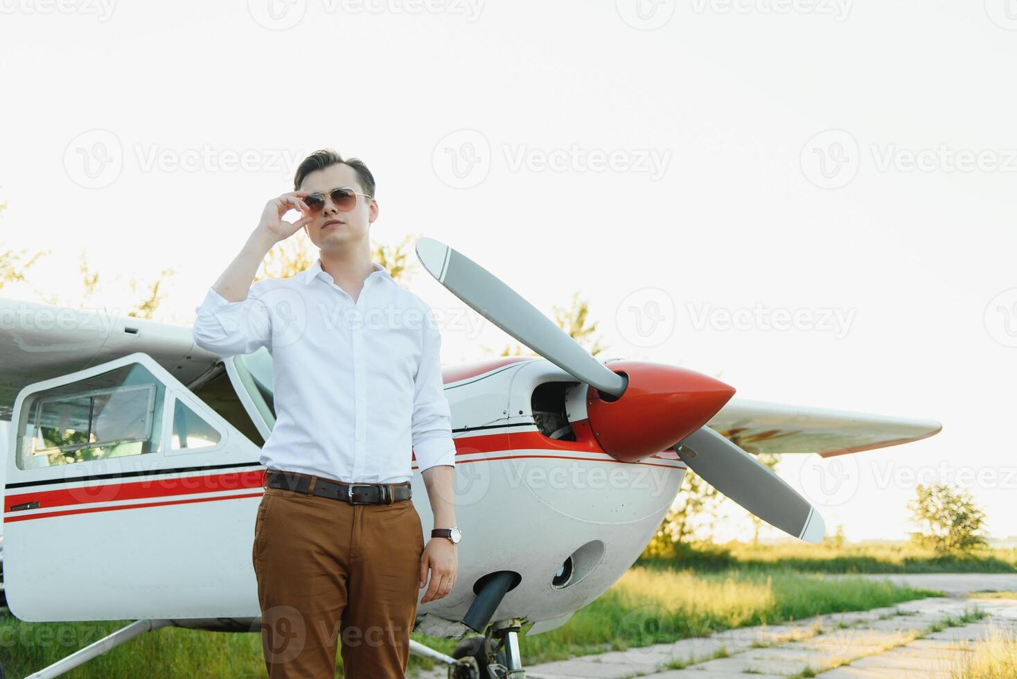 porträtt av stilig kommersiell pilot i kapten vit enhetlig stående och ser smart nära små privat helikopter på en landning punkt med privat jet terminal på internationell flygplats foto