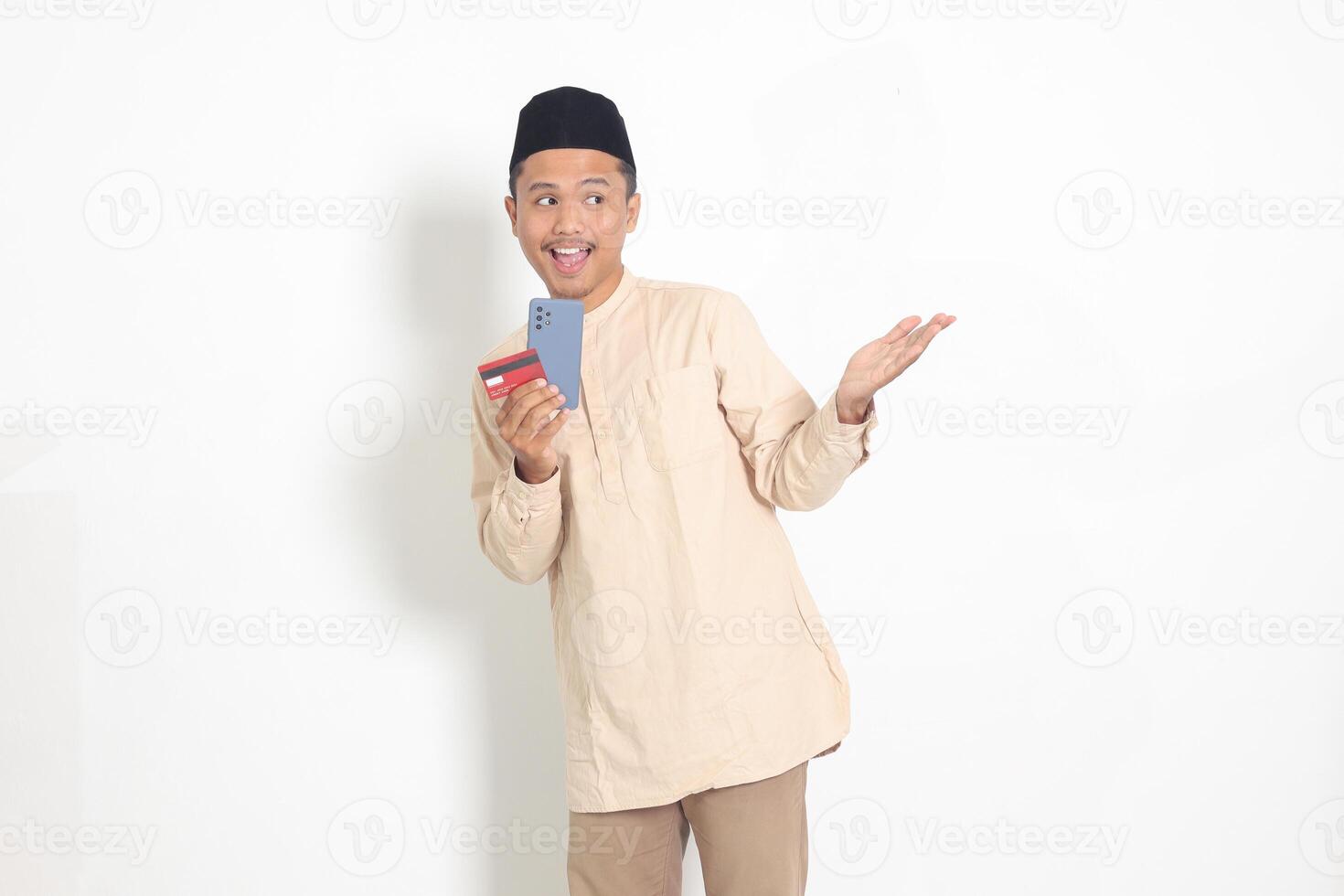 porträtt av attraktiv asiatisk muslim man i koko skjorta med kalott innehav en mobil telefon och kreditera kort medan pekande med finger till de sida. isolerat bild på vit bakgrund foto