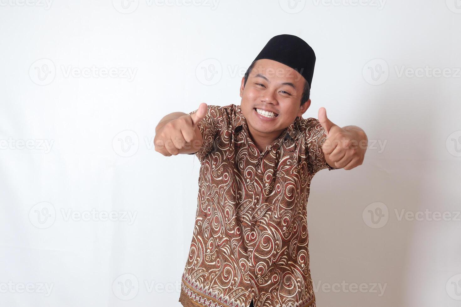 porträtt av upphetsad asiatisk man bär batik skjorta och Songkok leende och ser på kamera, framställning tummen upp hand gest. isolerat bild på grå bakgrund foto