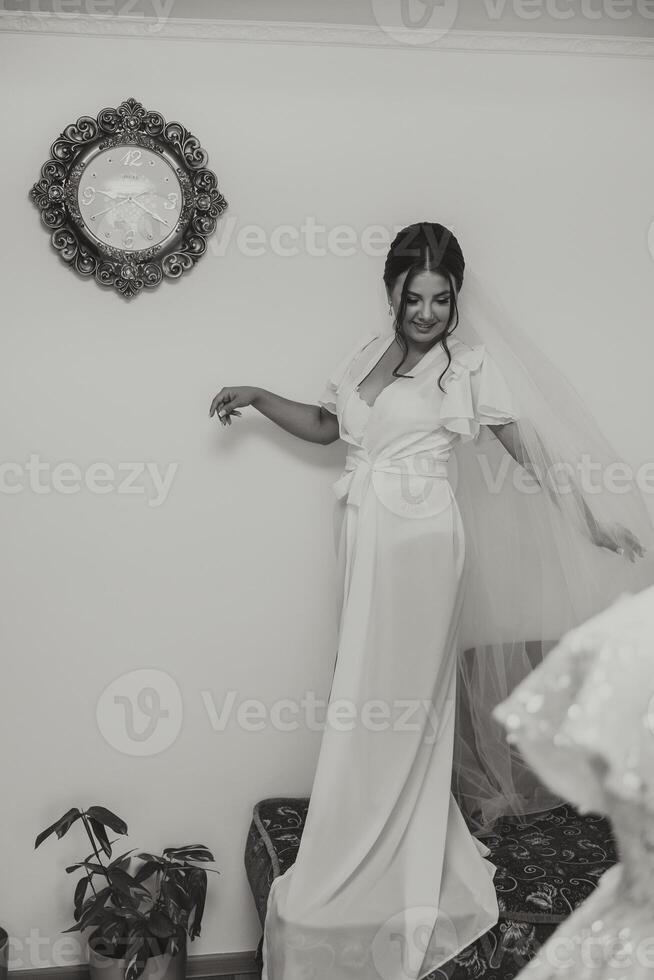 porträtt av ett otroligt skön flicka brud i en vit morgonrock i de sovrum och Framställ i de morgon- innan de bröllop ceremoni. de brud är stående förbi de vägg klocka foto