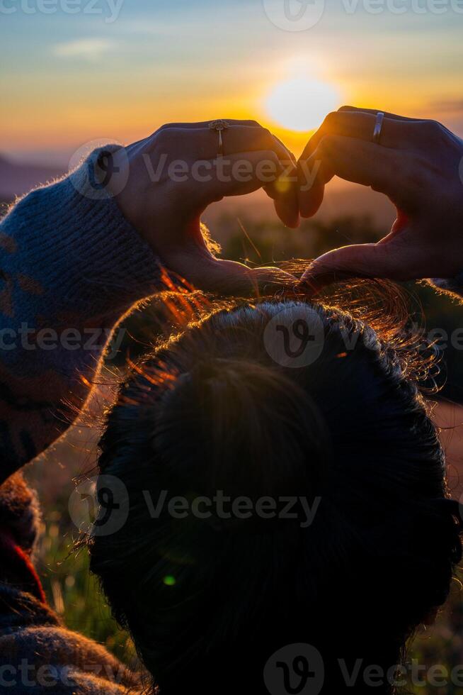 en ung flicka framställning hjärta symbol med henne händer på solnedgång foto