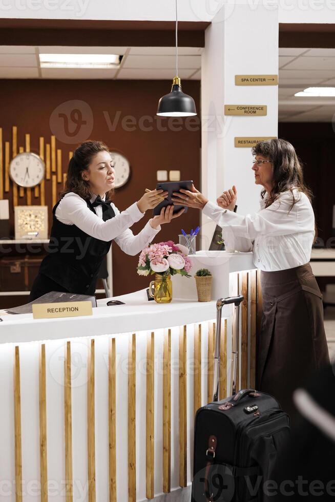 äldre kvinna ankommer på hotell reception som vänlig concierge assisterar med digital checka in form på läsplatta. gäst leende, tala. bekväm lobby med vardagsrum område. bagage redo för hotell rum. foto