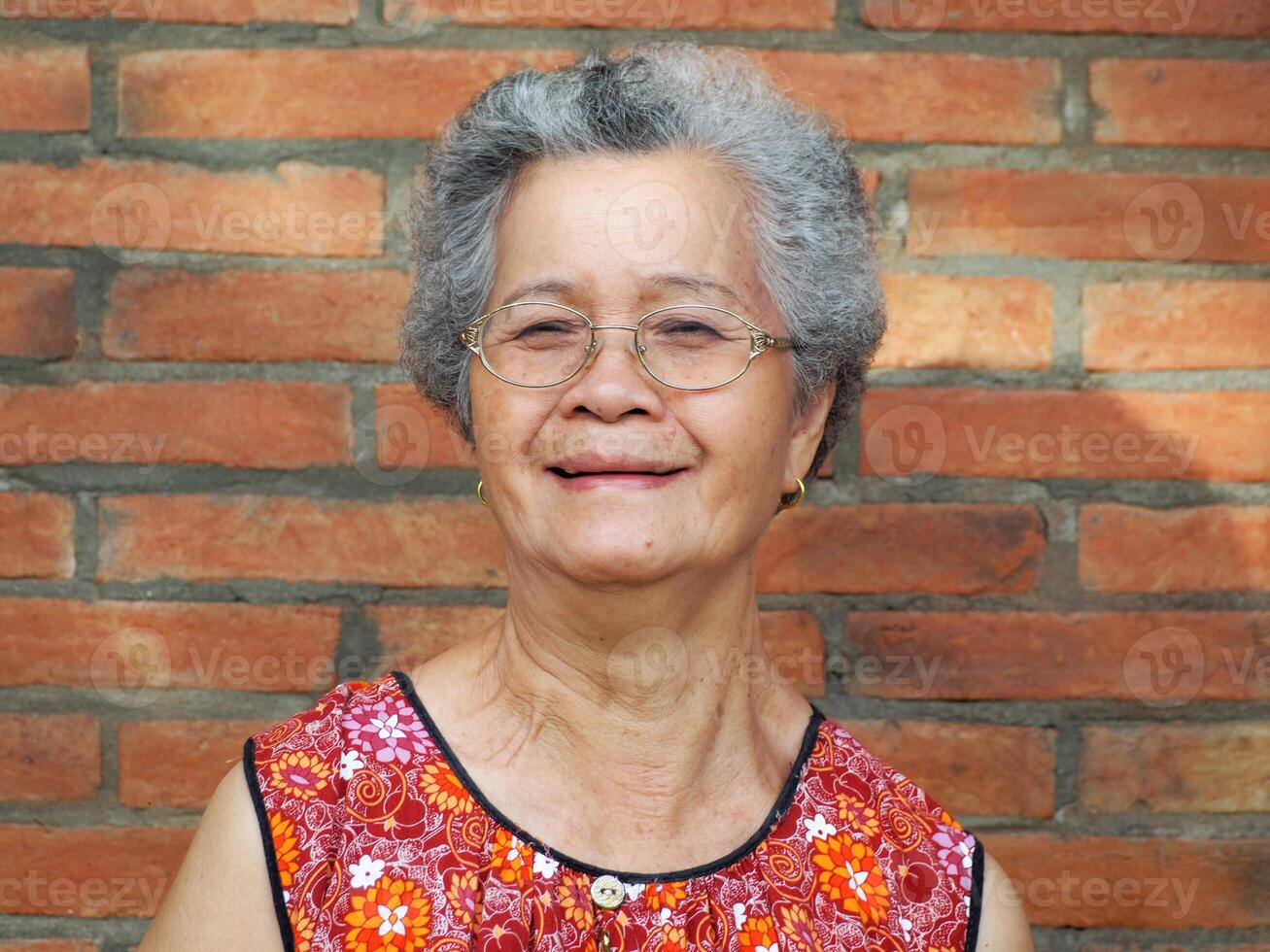 ett äldre asiatisk kvinna leende och ser på de kamera medan stående med en tegel vägg bakgrund. Plats för text. begrepp av åldrig människor och sjukvård foto