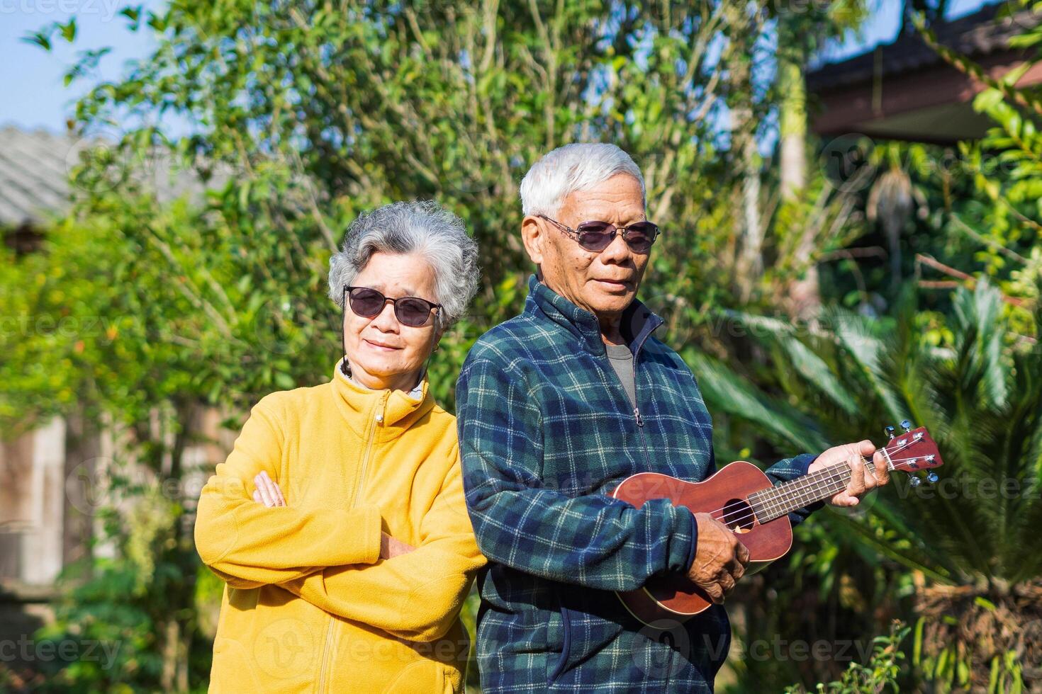 porträtt av en romantisk äldre asiatisk man spelar ukulele med hans fru medan stående i en trädgård. senior par leende och ser på de kamera. begrepp av åldrig människor och sjukvård foto