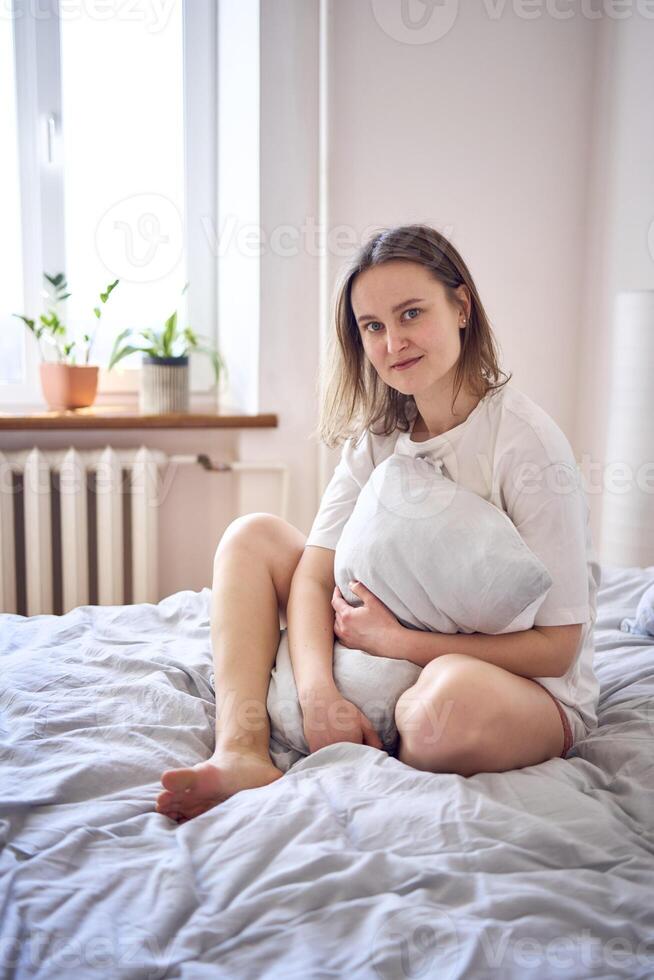 skön ung kvinna i vit t-shirt på säng kramas kudde foto