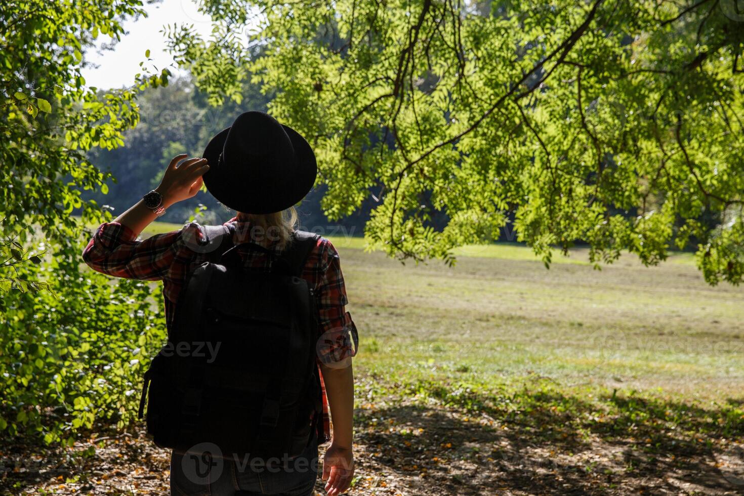 ung caucasian kvinna med en ryggsäck och hatt i de skugga av träd nära de fält. bakgrund, stänga upp. selektiv fokus.bakgrund suddig.hög kvalitet Foto