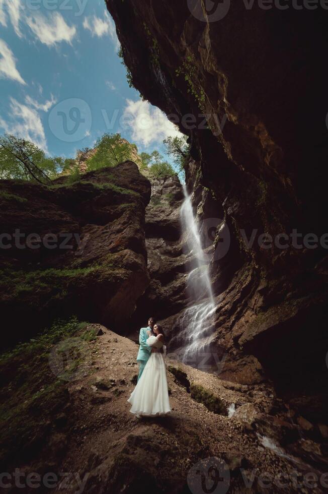 en ung man och hans fru är stående i ett omfamning i en kanjon mot de bakgrund av en hög vattenfall. nygifta bröllop par i de bergen foto