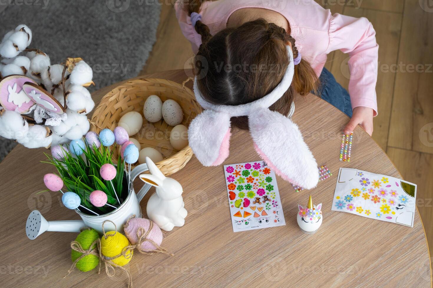 en söt flicka med rosa kanin öron gör ett påsk hantverk - dekorerar ett ägg i de form av en enhörning med strass, horn, blommor i de interiör av en hus med växter. foto