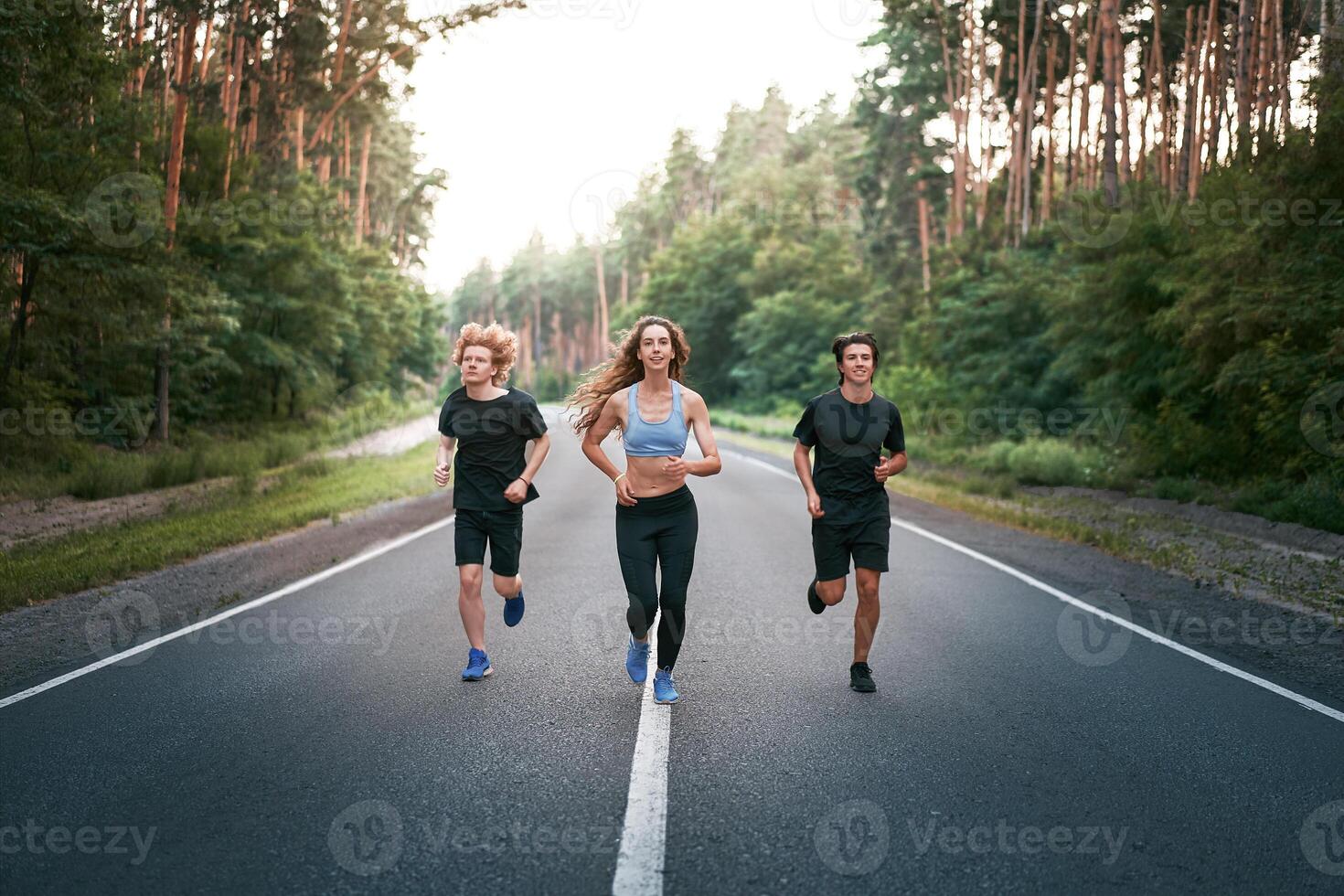 en grupp av tre människor idrottare ett flicka och två män springa på ett asfalt väg i en tall skog. foto