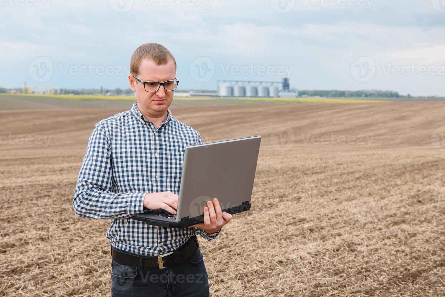 Lycklig jordbrukare med bärbar dator stående i vete fält i främre av spannmål silo foto