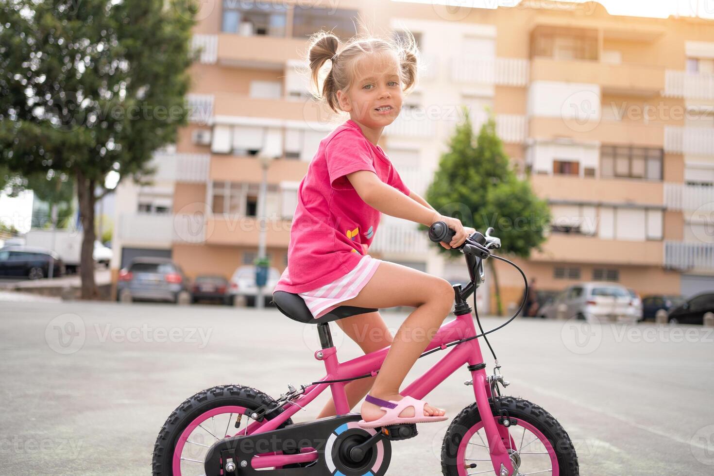 söt liten leende flicka ridning cykel cykel i stad på parkering solig sommar dag. aktiva familj fritid med ungar. foto