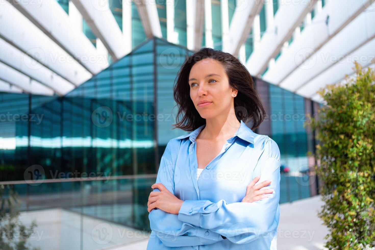 förtroende affärskvinna porträtt med korsade händer. Söt företag kvinna 30 år gammal stående nära kontor byggnad klädd blå skjorta. foto