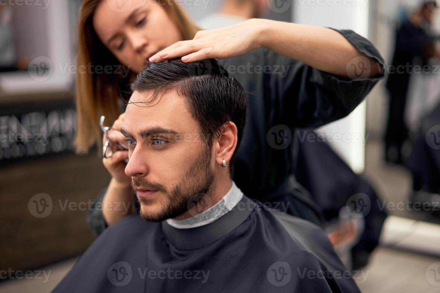 stilig blå ögon man Sammanträde i barberare affär. hårstylist frisör kvinna skärande hans hår. kvinna barberare. foto