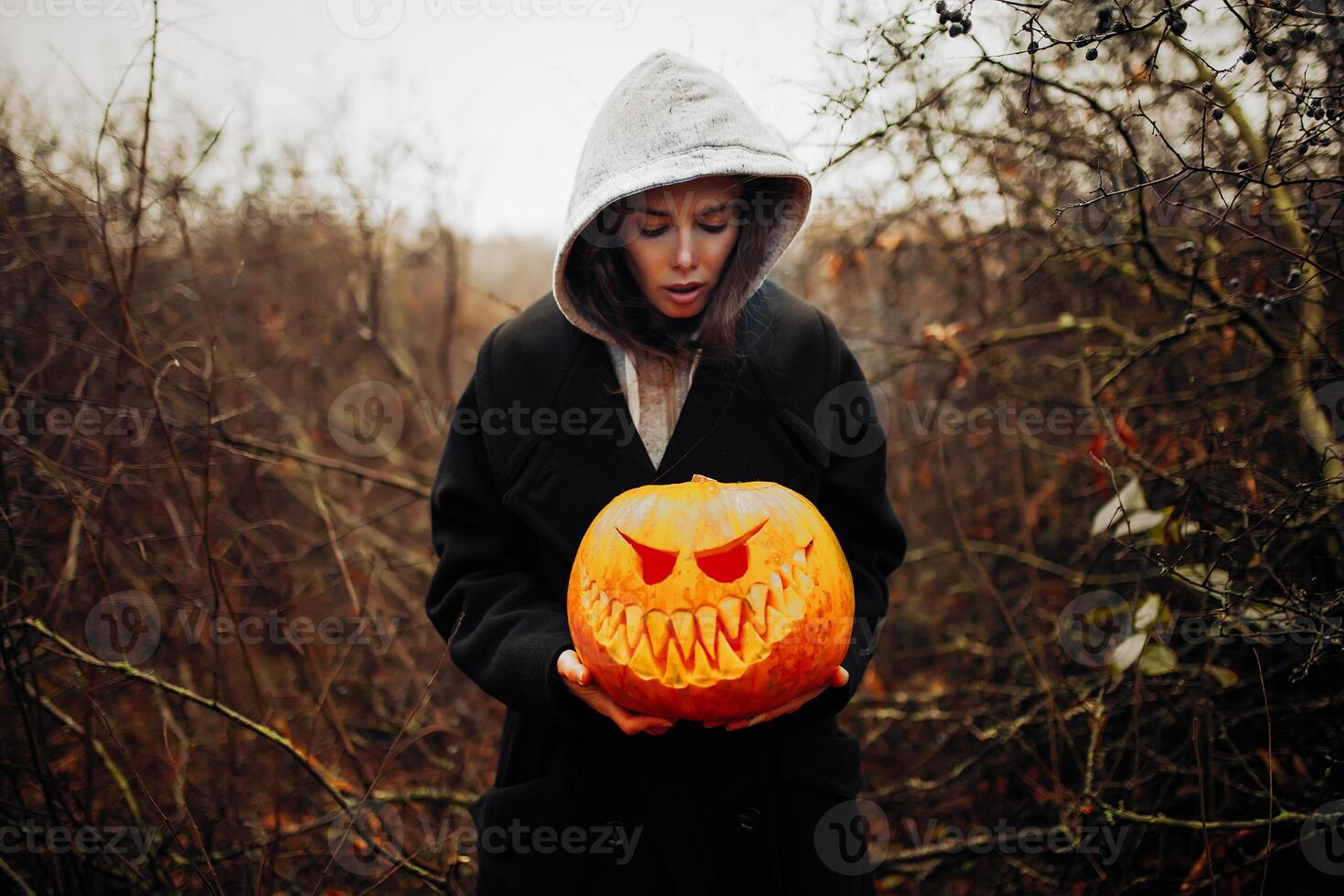 Lycklig halloween porträtt av svart modern livsstil häxa i huva och svart täcka mantel på trä- buske skog bakgrund. innehav pumpa i händer foto