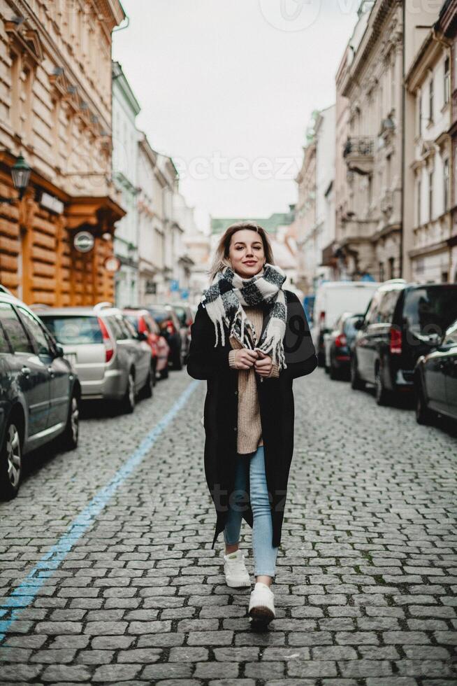 eleganta kvinna i svart täcka och scarf gående på kullersten gata. urban mode, stad livsstil begrepp - fotgängare i stadsbild med parkerad bilar, vinter- scen, urban utforskning och modern foto