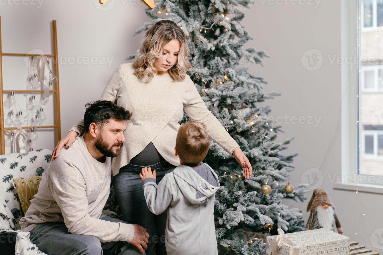 jul familj lycka porträtt av pappa, gravid mamma och liten son Sammanträde fåtölj på Hem nära jul träd kram leende foto