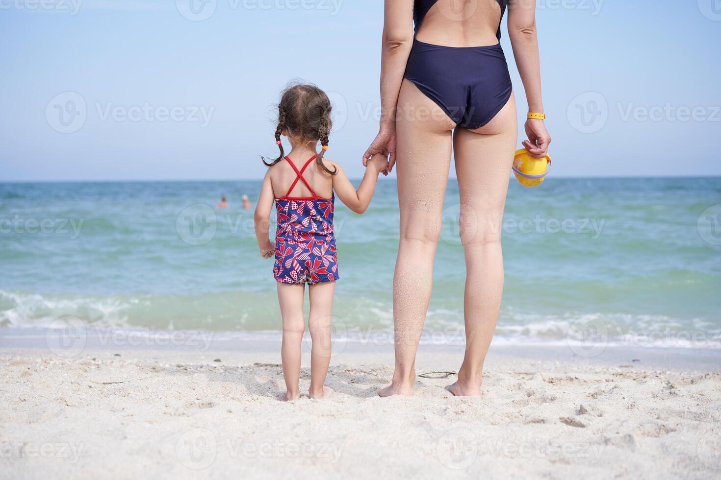 mor dotter strand tillsammans bak- se oigenkännlig caucasian kvinna liten flicka badkläder stående havet tillbaka. foto