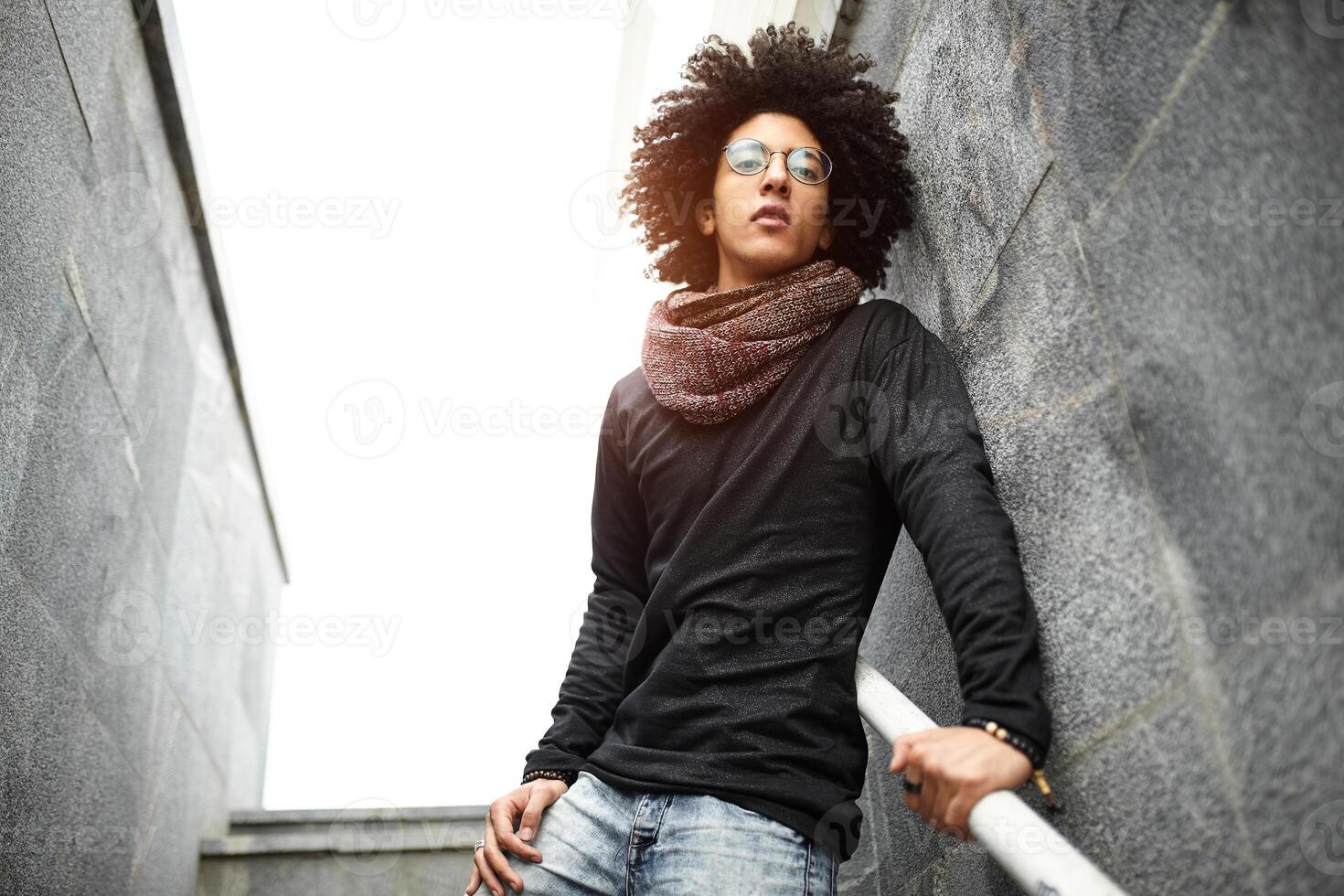 stilig ung man av blandad lopp med en frisyr i modern kläder, jeans och en scarf Framställ mot de bakgrund av en vägg och trappa tillverkad av keramisk granit plattor foto