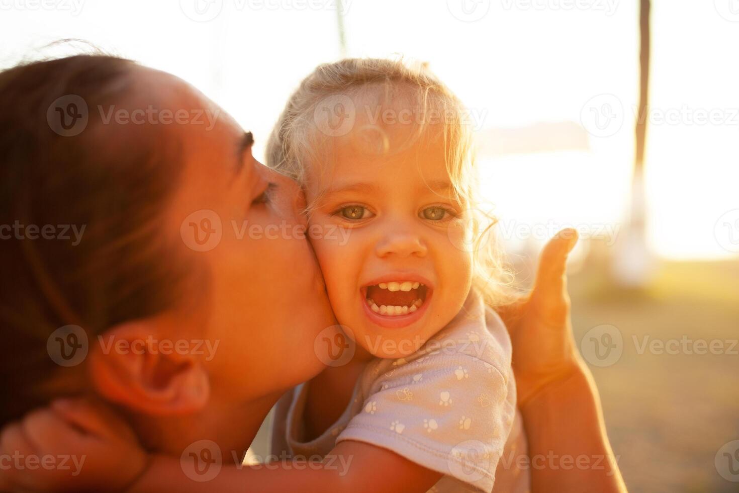 en stor porträtt, mor kyssar henne dotter i de strålar av de miljö Sol. familj värden, Lycklig barn foto