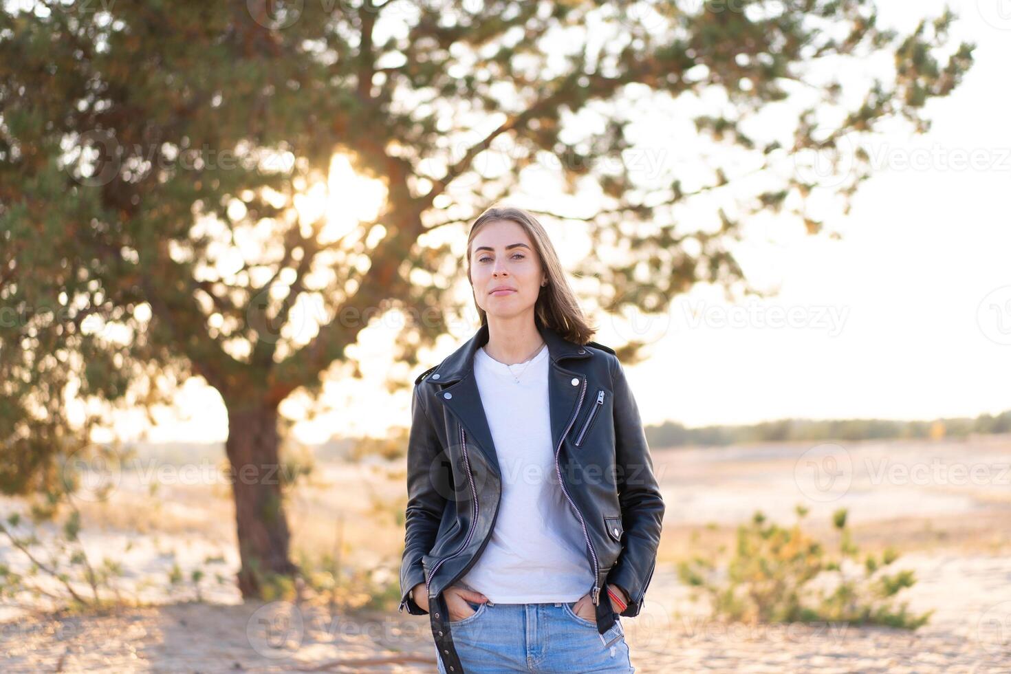 ung skön caucasian kvinna klädd i en läder jacka och jeans står på de strand på en trä bakgrund. foto