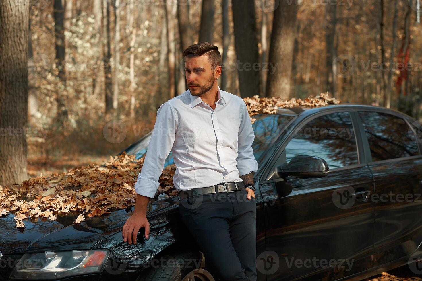 en stilig ung caucasian man bär företag kostym stående nära svart bil på natur. foto