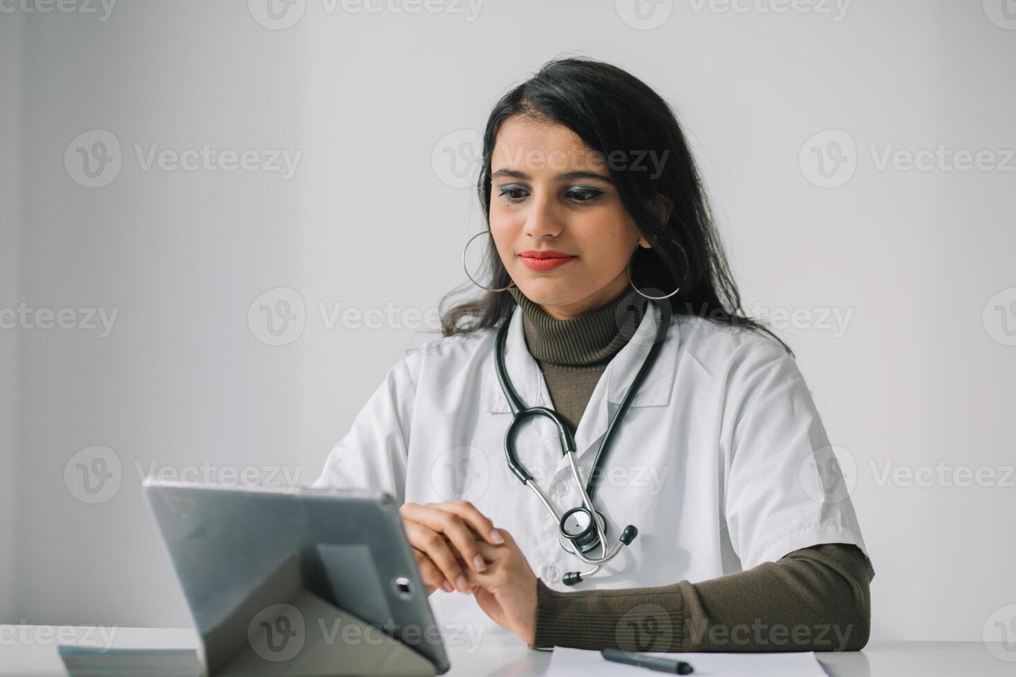 indisk kvinna läkare i en vit täcka med en stetoskop utför ett uppkopplad samråd i henne kontor. kvinna läkare kommunicerar med de visare direkt in i de kamera foto