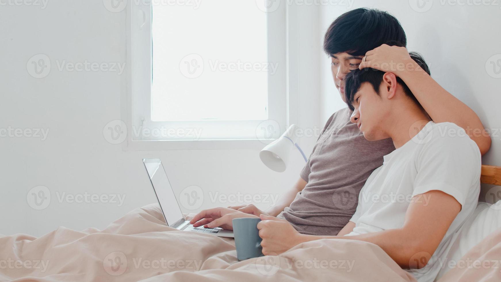Asiatiska homosexuella män par använder dator bärbar dator och dricker kaffe i moderna hem. ung asien älskare man glad slappna av vila tillsammans efter att ha vaknat, titta på film liggande på sängen i sovrummet hemma på morgonen foto