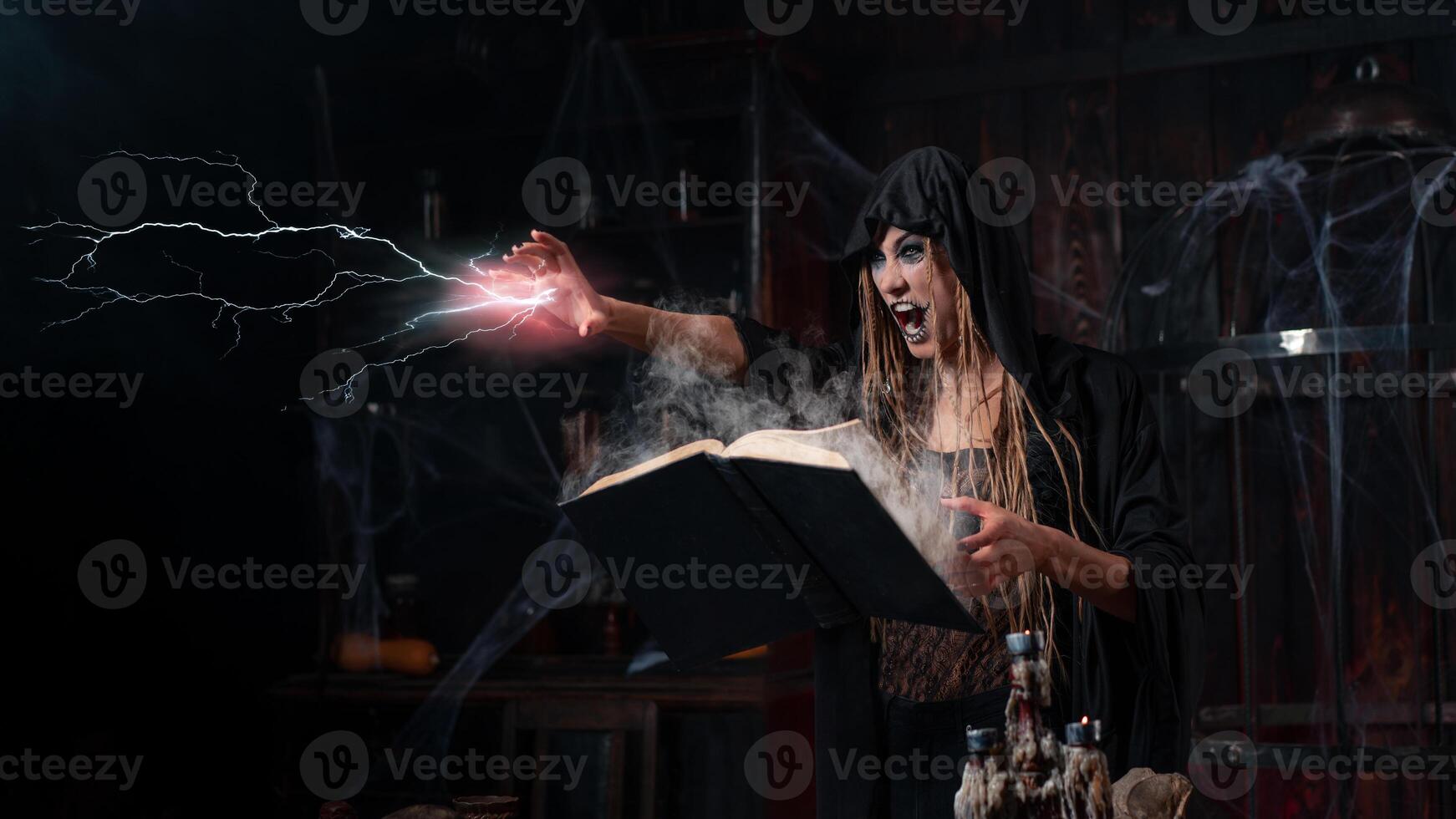 halloween begrepp. häxa klädd svart huva med dreadlocks stående mörk hålan rum använda sig av magi bok för framkallning magi stava foto