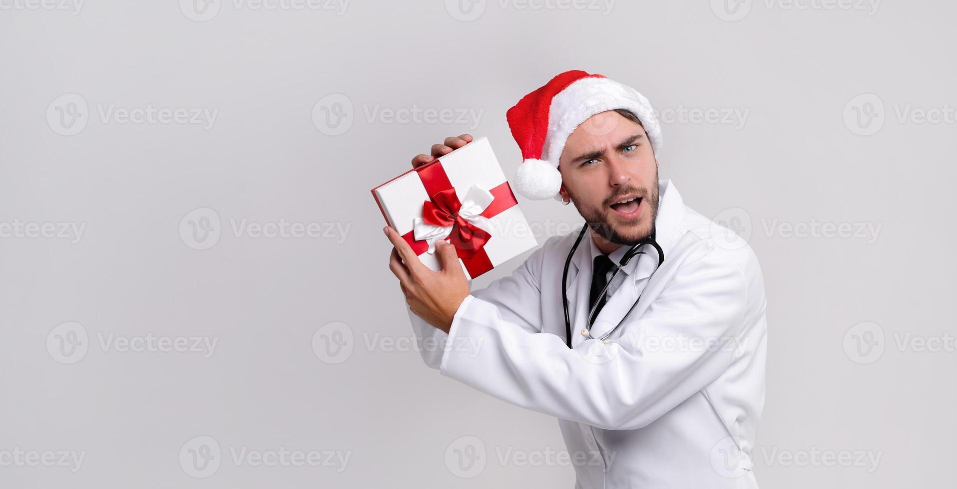 ung stilig läkare i vit uniforme och santa claus hatt stående i studio på vit bakgrund leende och finger i kamera foto