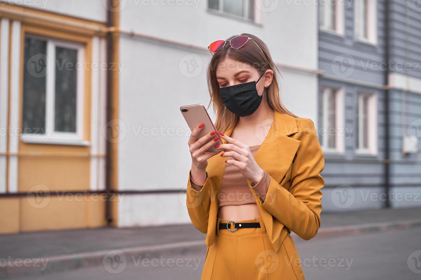 ung skön caucasian flicka 20 år ha på sig svart ansikte mask skydd mot epidemi coronavirus covid-19 promenader ner de gata utseende på de telefon skärm. foto