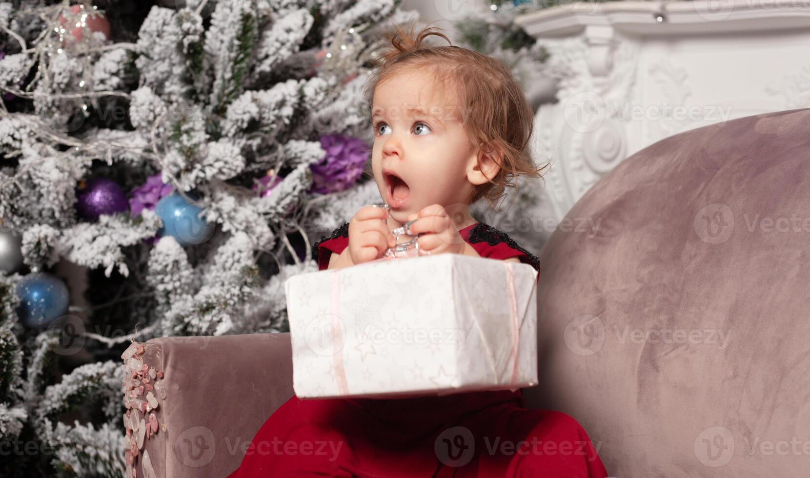 en skön söt liten flicka klädd i ett elegant kväll röd klänning sitter på de soffa och öppnas en ny år gåva. med jul träd på de bakgrund foto