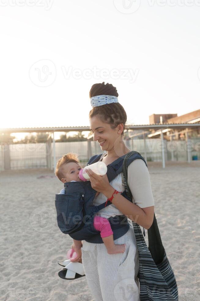 en små barn sitter i en ryggsäck och promenader längs med de mor längs de stranden. sommar familj semester begrepp foto