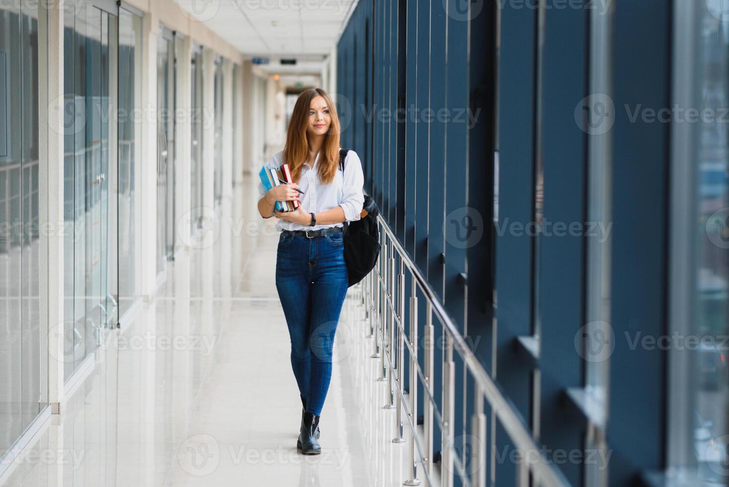 glad brunett studerande flicka med svart ryggsäck innehar böcker i modern byggnad. kvinna studerande stående med böcker i högskola hall foto