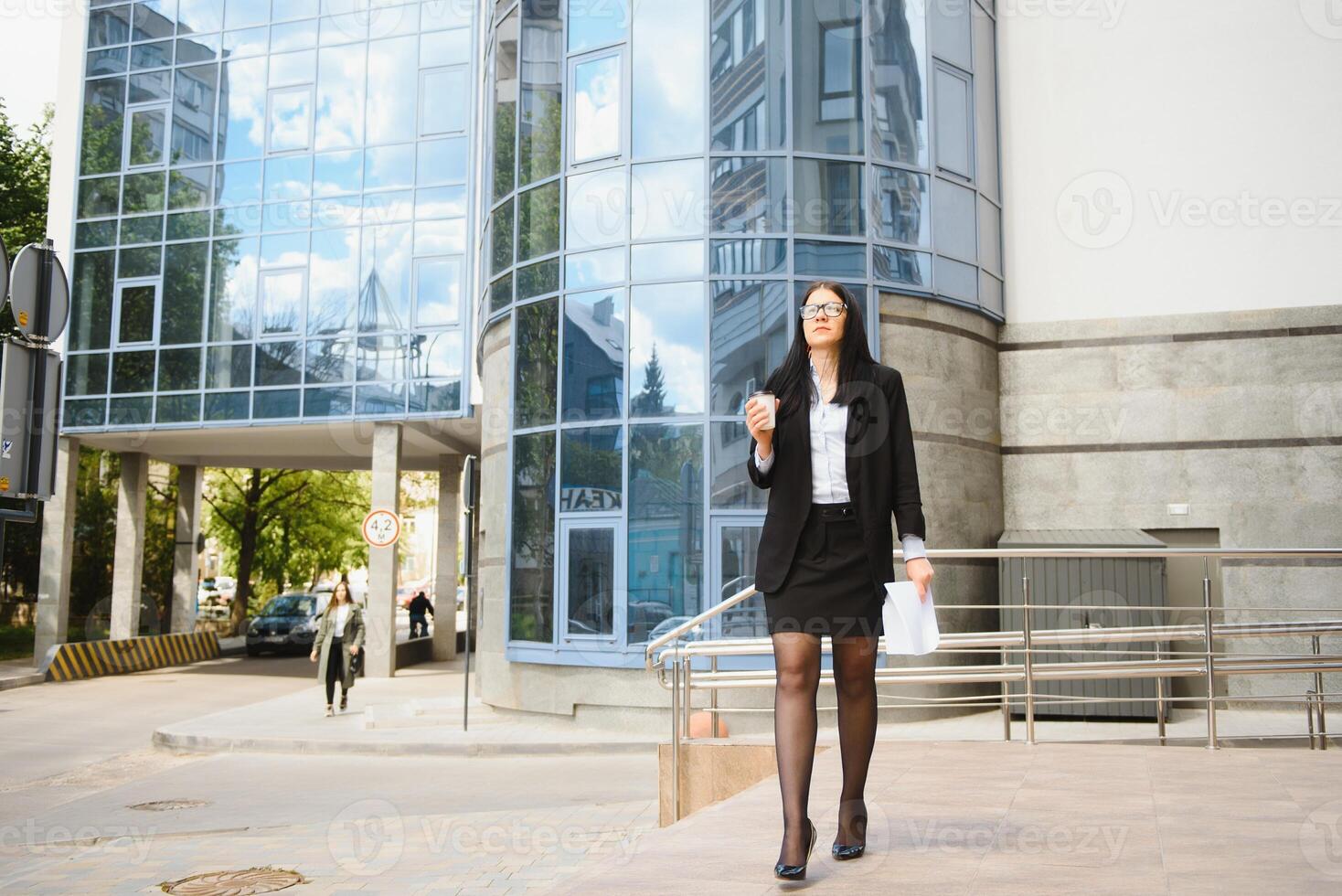 ung flicka kontor arbetstagare i glasögon, mörk jacka och blå skjorta vistelser på stad gata nära kontor byggnad och innehar henne dokument. foto