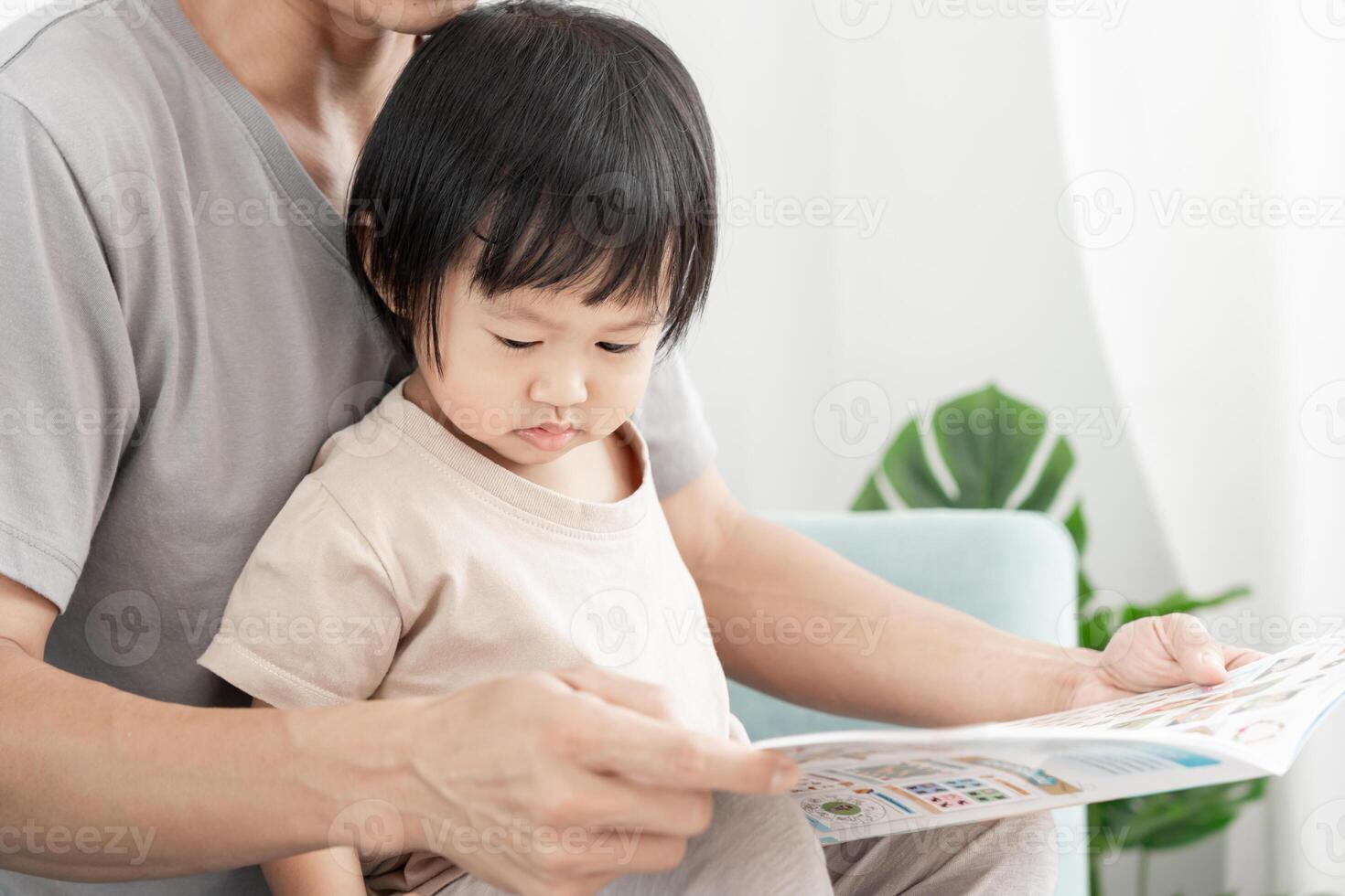 Lycklig asiatisk far koppla av och läsa bok med bebis tid tillsammans på Hem. förälder sitta på soffa med dotter och läsning en berättelse. lära sig utveckling, barnomsorg, skrattande, utbildning, berättande, öva. foto