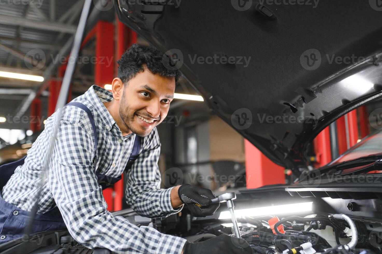 indisk bil mekaniker stående och arbetssätt i service station. bil specialister granskning de lyft bil. professionell reparatörer bär mekaniker enhetlig i blå Färg. foto