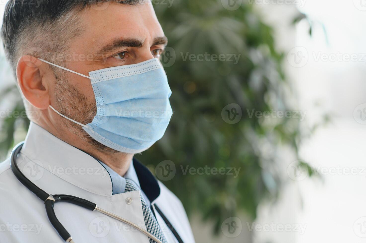 mogna gammal medicinsk sjukvård professionell läkare bär vit täcka, stetoskop, glasögon och ansikte mask stående i sjukhus. medicinsk personal hälsa vård skydd begrepp. porträtt foto